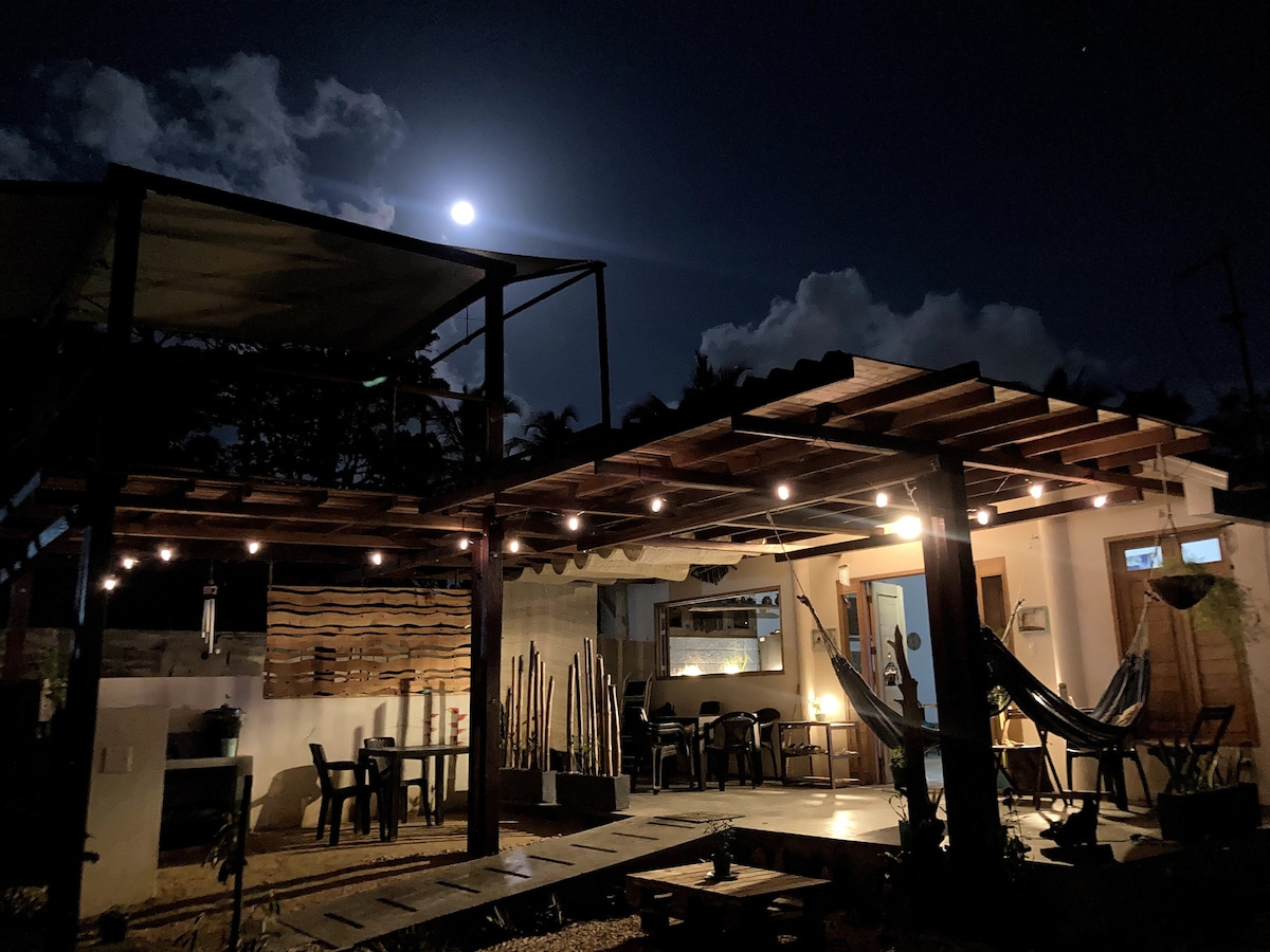 A spacious outdoor area features a wooden structure adorned with string lights, complementing the warm glow from nearby indoor lighting. A hammock and several chairs create a comfortable setting underneath the night sky, with a full moon illuminating the scene.