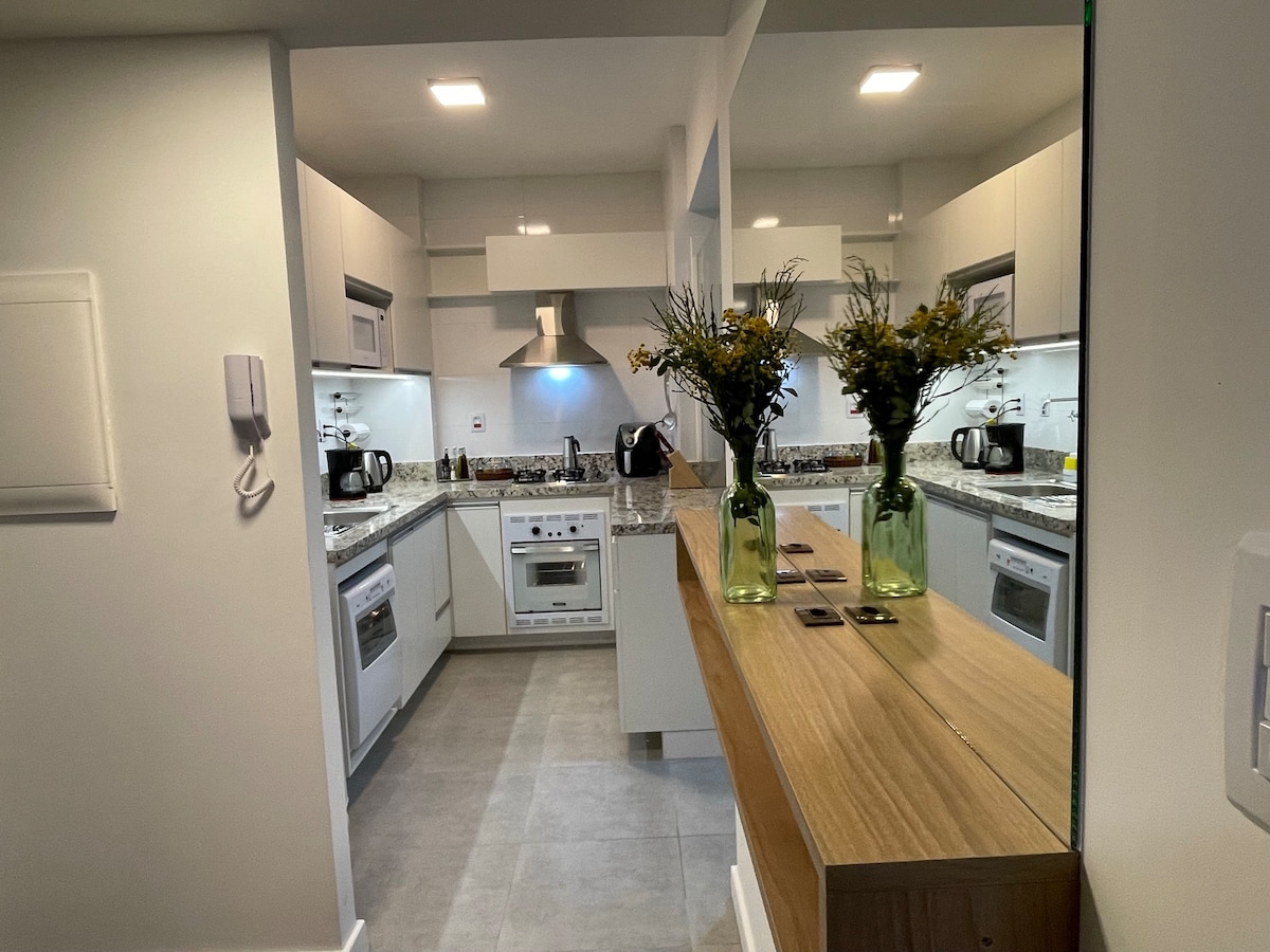 The kitchen area features modern cabinetry and a spacious countertop, highlighted by a stylish wooden table. Fresh flowers in green vases bring a touch of color to the room. The appliances, including a stove and microwave, are neatly integrated into the design.