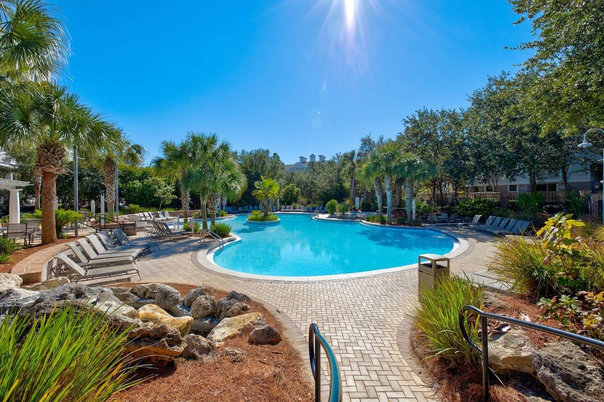 A spacious pool area is surrounded by palm trees and lush landscaping. Sun loungers are positioned around the pool, which gleams under a clear blue sky. The pathway features patterned stonework, leading to vibrant greenery and decorative rocks, creating a serene environment.
