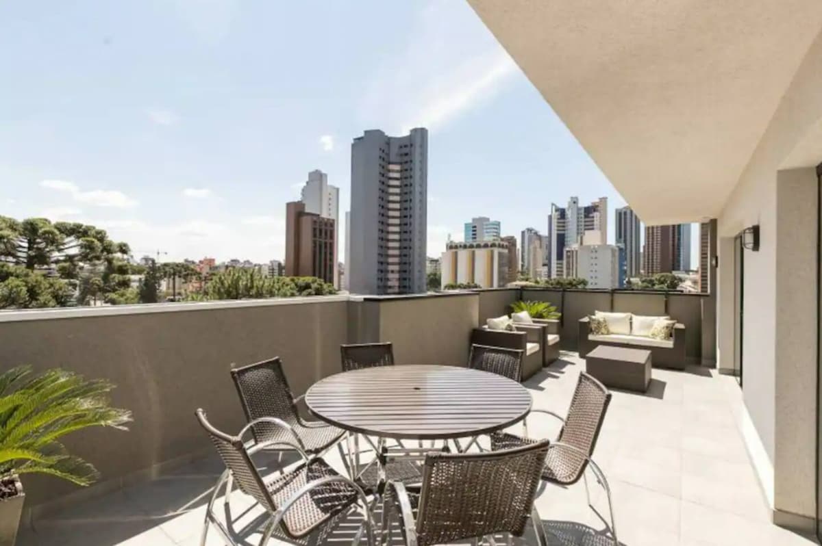 An outdoor terrace features a round table surrounded by six comfortable chairs. Lush green plants decorate the space, while the backdrop showcases a panoramic view of a city skyline with a mix of tall buildings under a bright blue sky.