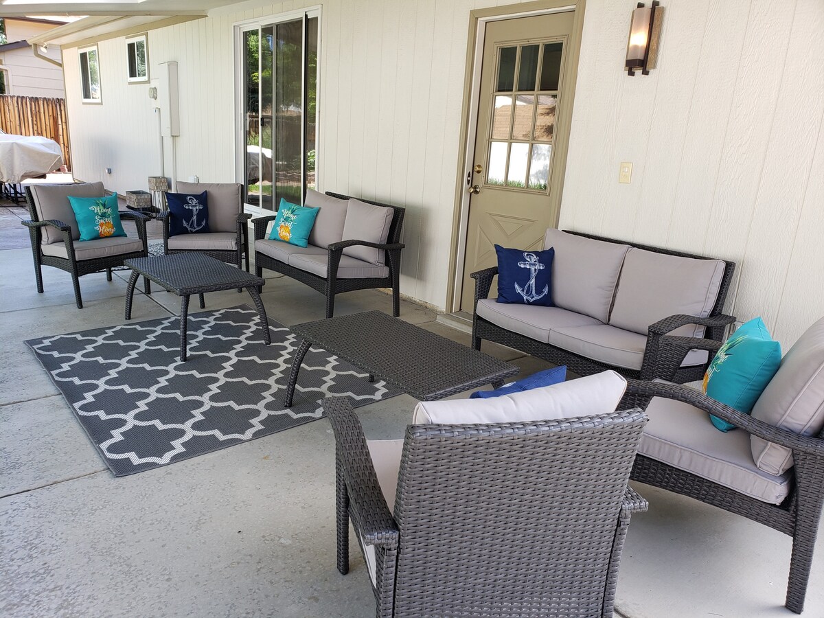 An outdoor seating area features a variety of wicker furniture arranged around a decorative area rug. Cushions in shades of blue and green accent the seating. Large windows and a door provide access to the interior space, enhancing natural light and openness.