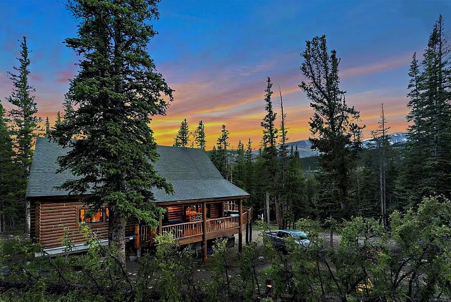 A log cabin is nestled among tall pine trees, with a wraparound porch visible. The warm glow from the windows suggests a cozy interior. A picturesque sunset illuminates the sky in shades of orange and pink, providing a peaceful mountain backdrop.