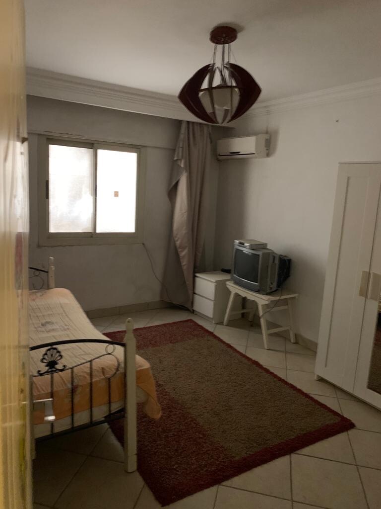 A private room is presented, featuring a single bed with a light-colored sheet. A small television is situated on a stand, alongside a white bedside table. The room includes a closet and is illuminated by a ceiling light fixture. A window with light curtains allows natural light.