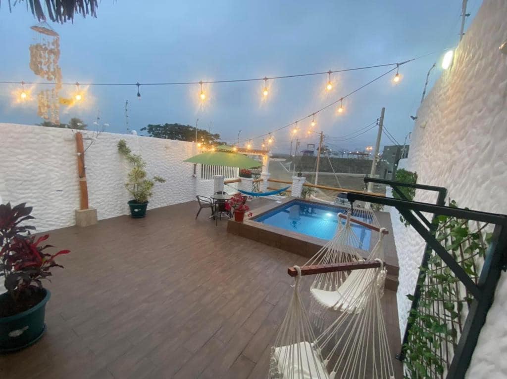 A tranquil outdoor area features a small swimming pool surrounded by wooden decking and lush plants. Soft lighting from hanging bulbs enhances the space, which includes a hammock and seating for relaxation. The walls are textured and painted white, creating a serene atmosphere.