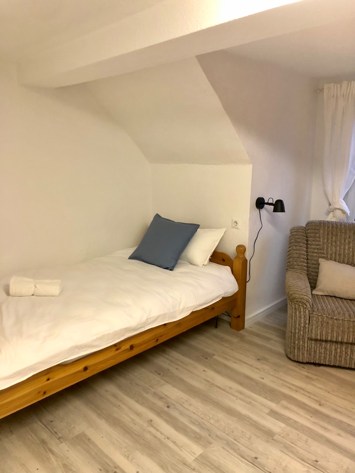 A simple sleeping area features a wooden bed with white linens and a blue accent pillow. A comfortable chair is positioned nearby, with a wall-mounted light providing soft illumination. Light-colored flooring complements the bright, airy atmosphere.