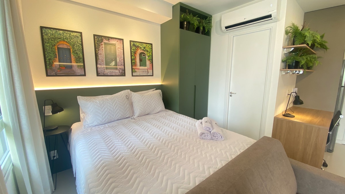 A modern bedroom is featured, with a spacious bed adorned in crisp white linens. Green walls are complemented by artwork with leafy exteriors. Soft lighting enhances the calming ambiance, while potted plants are arranged above a stylish desk. A small sofa is situated at the foot of the bed.