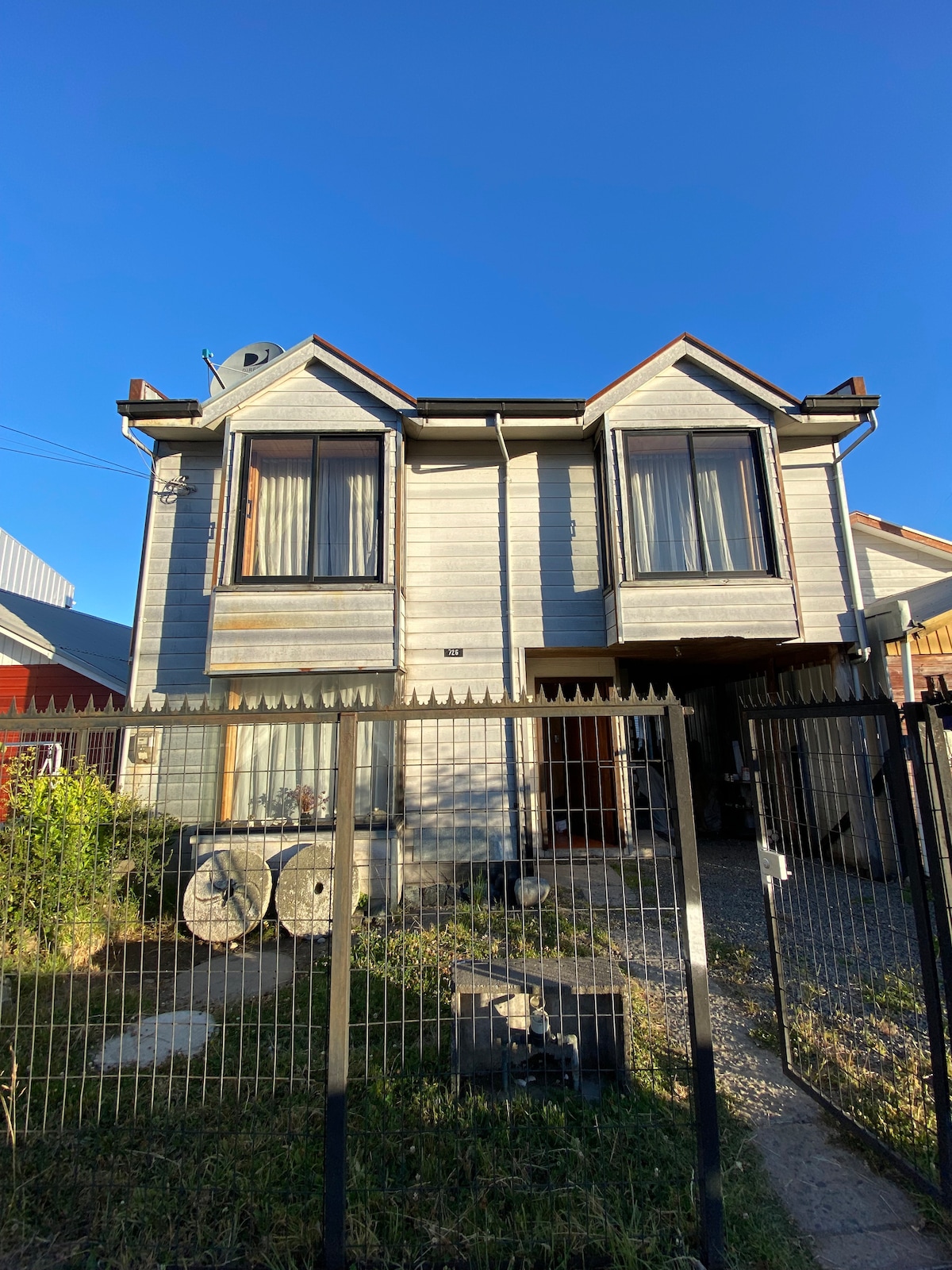 The exterior of the house features a two-story wooden structure with large windows adorned by long curtains. A secure fence surrounds the property, with a small garden visible in front. The clear blue sky creates a bright backdrop for the home.