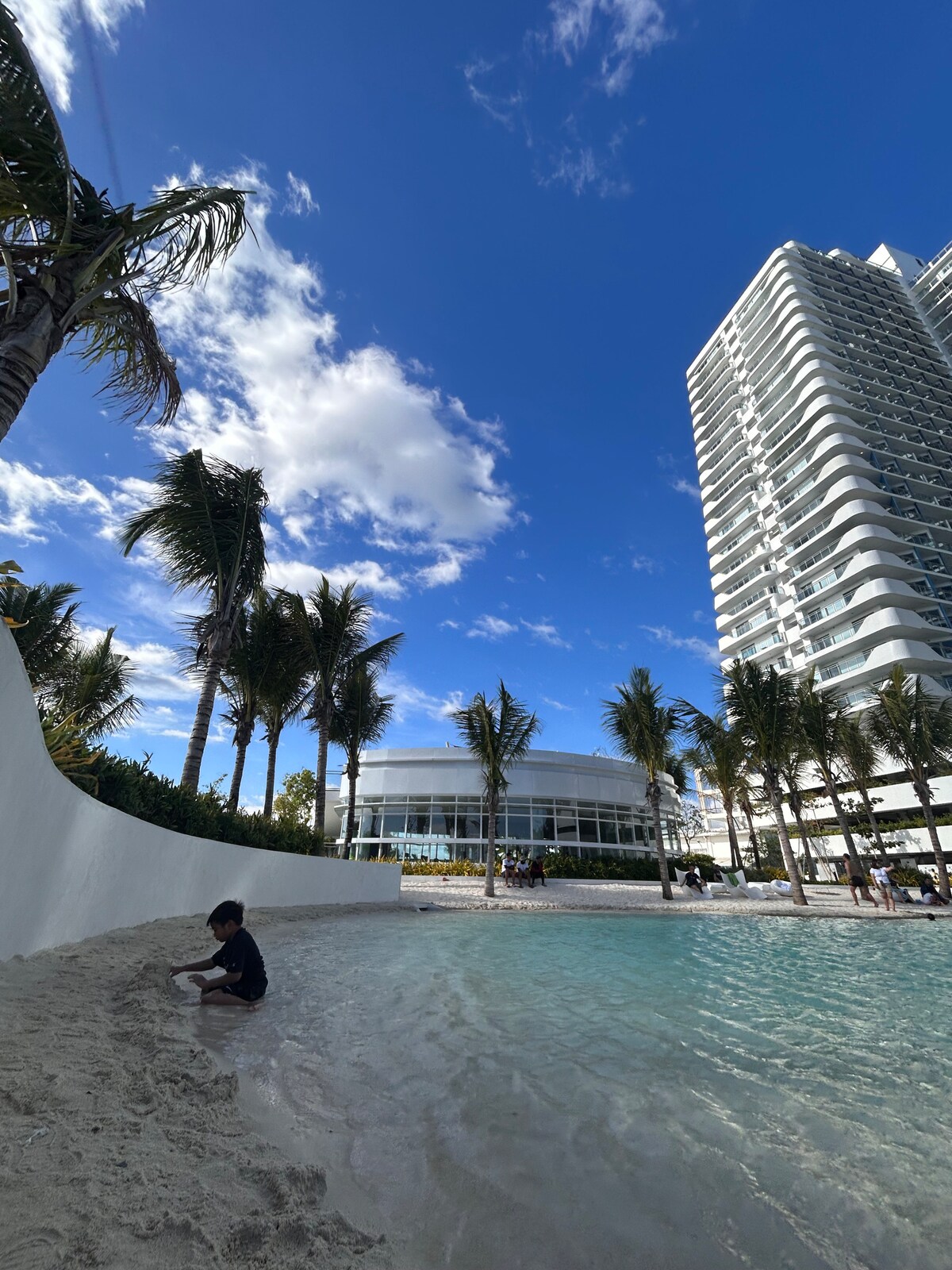 A serene scene is captured at the manmade beach and wave pool, featuring a child playing in the soft sand near calm waters. Towering palm trees sway gently in the breeze, while the modern building structure reflects the blue sky, creating a relaxed atmosphere.