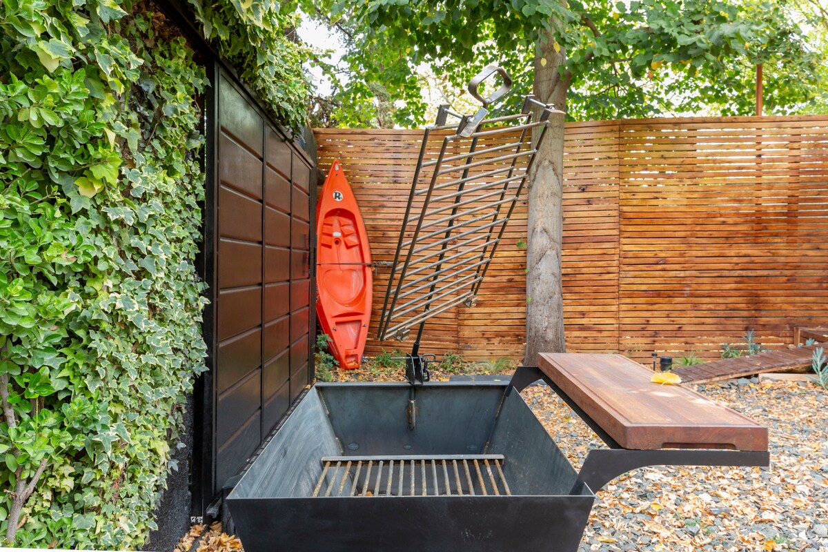An outdoor grilling area features a charcoal grill, with a wooden countertop alongside. A bright orange kayak leans against a wooden fence, surrounded by lush greenery and gravel landscaping.