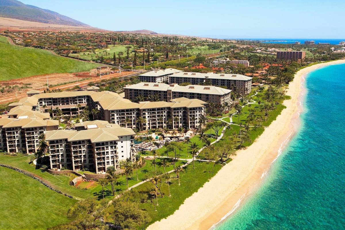 An aerial view of the Westin Kaanapali Ocean Resort Villas showcases a sprawling beachfront property surrounded by lush greenery. The resort buildings are positioned closely to the sandy beach, with crystal blue waters nearby. Mountains and hills are visible in the background, completing the tranquil coastal scene.