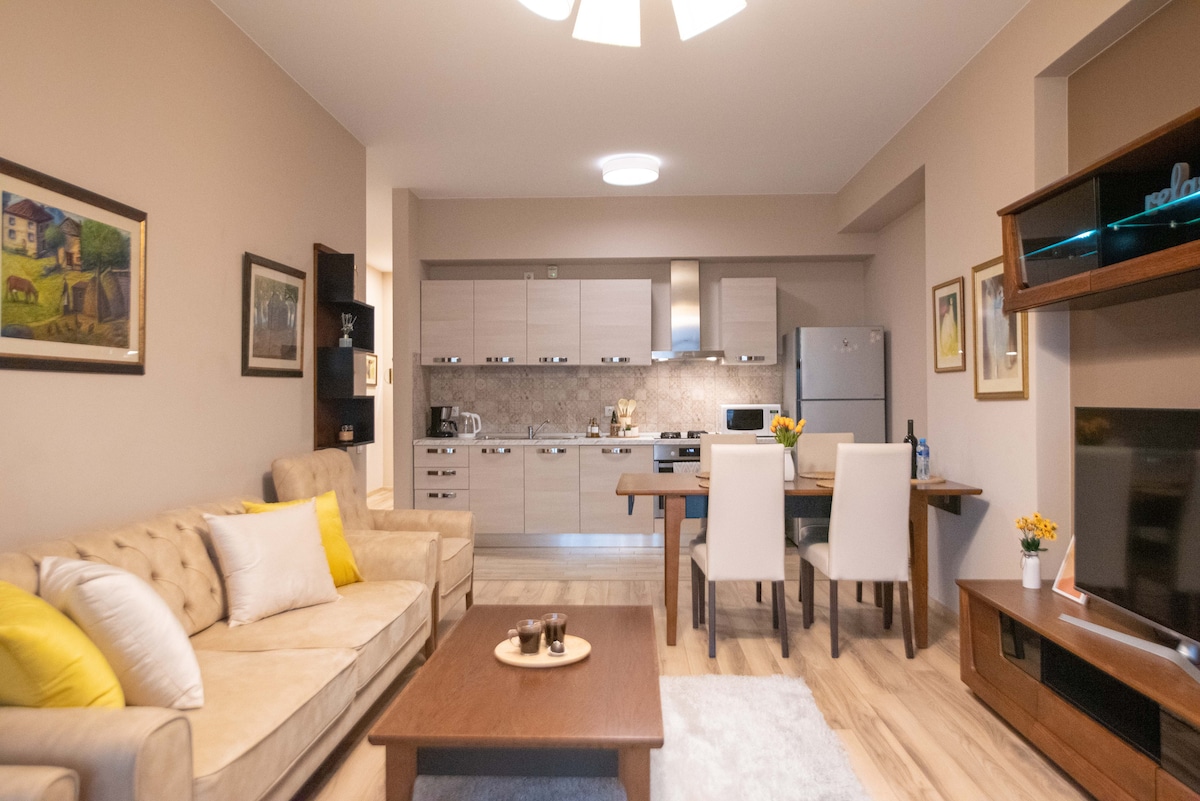 A spacious living area features a comfortable sofa and armchairs in a light beige hue, complemented by decorative pillows. A modern kitchen, equipped with appliances and a dining table for four, is visible. Soft light illuminates the room, enhancing the neutral color palette.