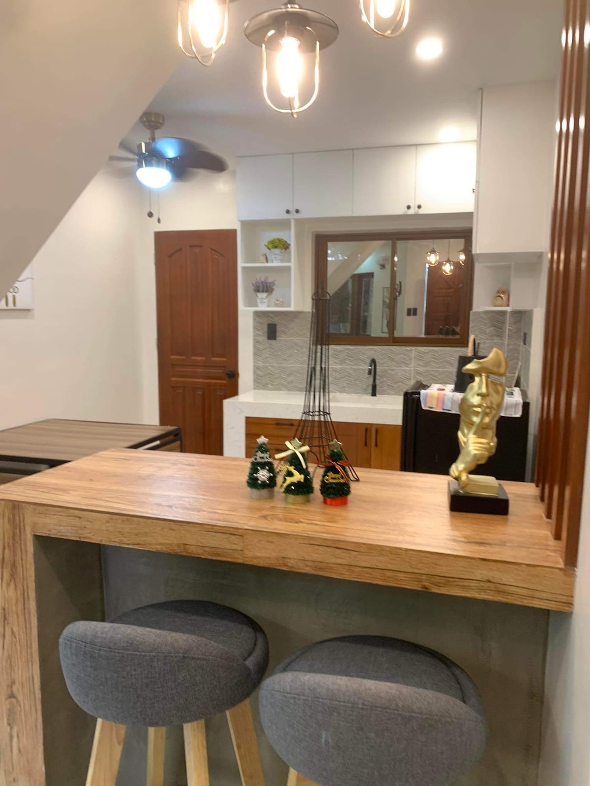 A stylish kitchen nook features a wooden bar with two grey stools. Decorative items, including small figurines and festive decorations, are placed on the bar. The space is brightened by stylish pendant lighting and a ceiling fan, contributing to a cozy atmosphere.