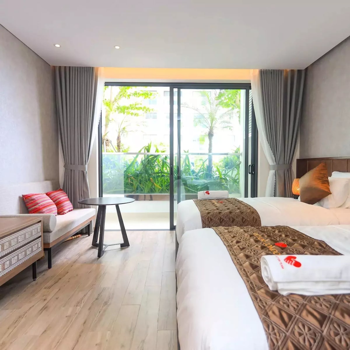 A modern hotel room is presented with two spacious beds, complemented by decorative throws. A small table is positioned between the beds, and floor-to-ceiling sliding glass doors provide views of greenery outside. Natural light brightens the room through the curtains.