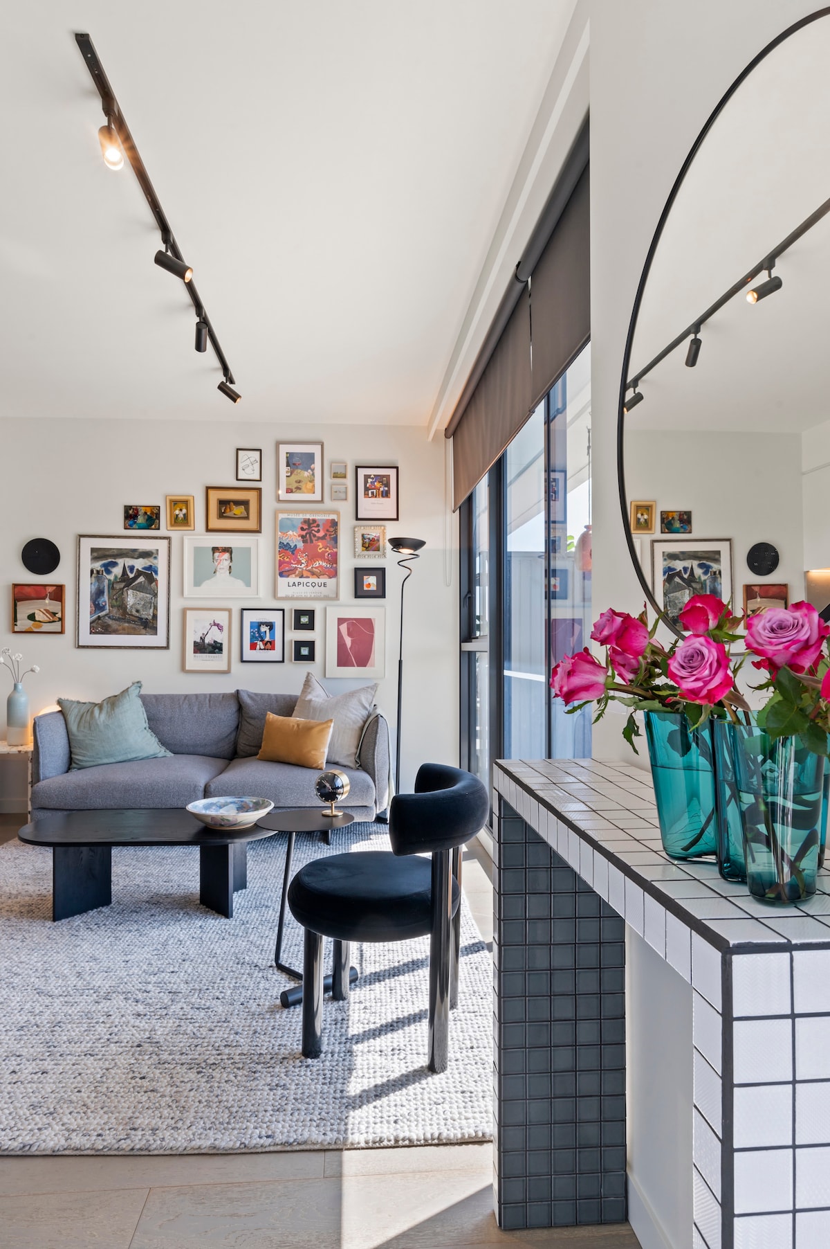 A modern living area features a comfortable couch adorned with decorative cushions, complemented by a round table. A gallery wall showcases an assortment of framed artwork. A mirror reflects midday light while a vase of fresh roses adds a touch of color to the space.