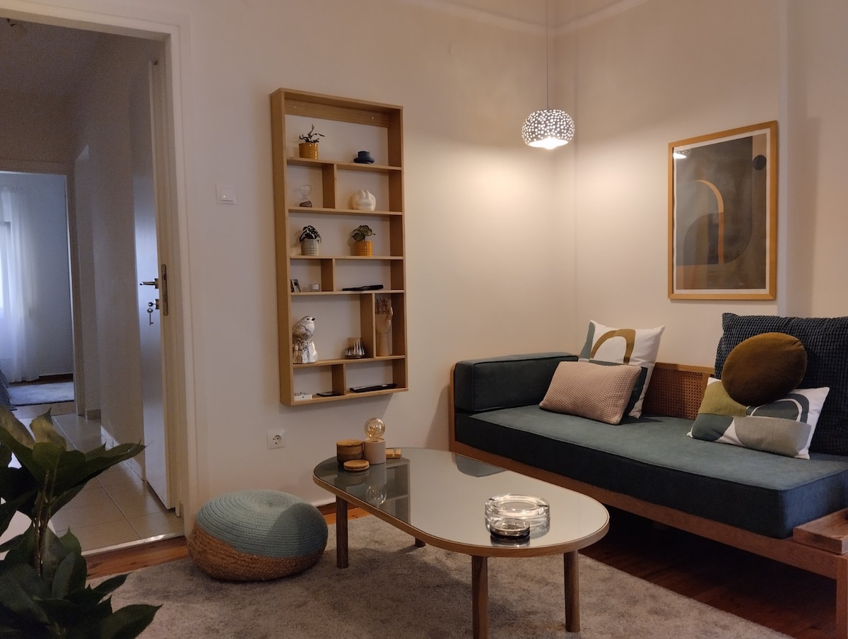 A well-arranged seating area features a dark green couch adorned with cushions in various textures. A round coffee table sits in front, topped with decorative items. Soft lighting from a modern fixture accentuates the neutral walls and a shelf displaying curated decor.