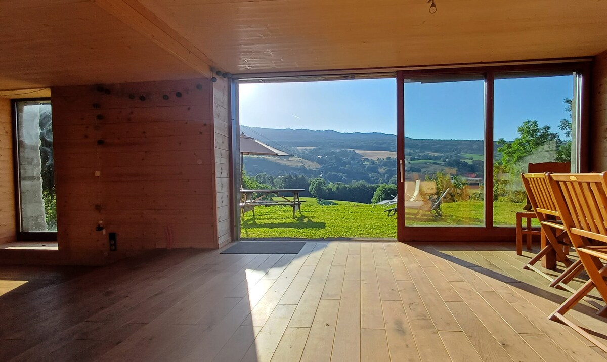 A spacious living area features large glass doors that open to a panoramic view of the surrounding landscape. Wooden chairs are arranged around a dining table, allowing for comfortable seating. Natural light floods the space, enhancing the warm wooden interior.