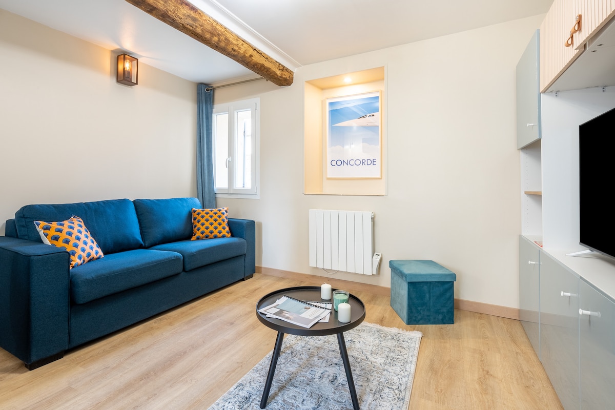 A cozy living area features a comfortable blue sofa adorned with decorative pillows. A low coffee table holds magazines and a candle. A rounded pouf adds extra seating, while a wall-mounted TV and a framed artwork enhance the space's modern design. Natural light enters through a window.