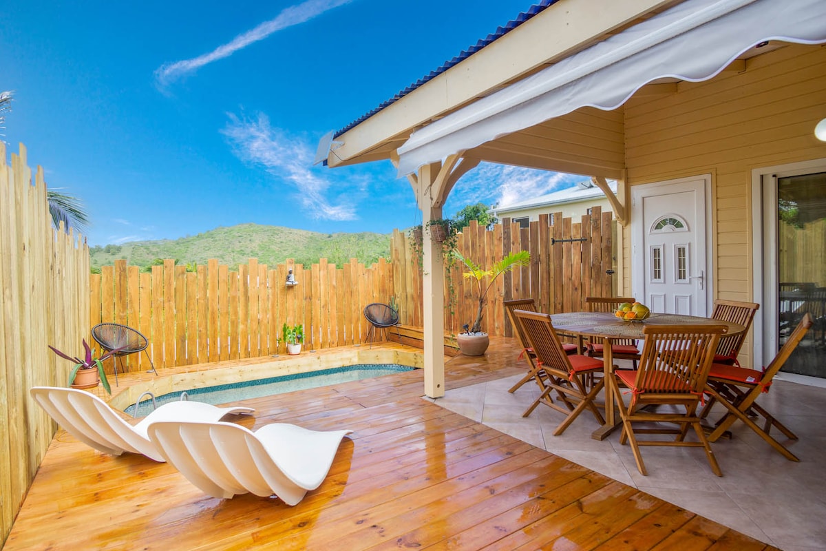 A welcoming outdoor dining area features a wooden table surrounded by matching chairs, set against a backdrop of a wooden fence. A small pool is visible, with lounge chairs placed nearby. Bright blue skies and green hills can be seen in the distance.