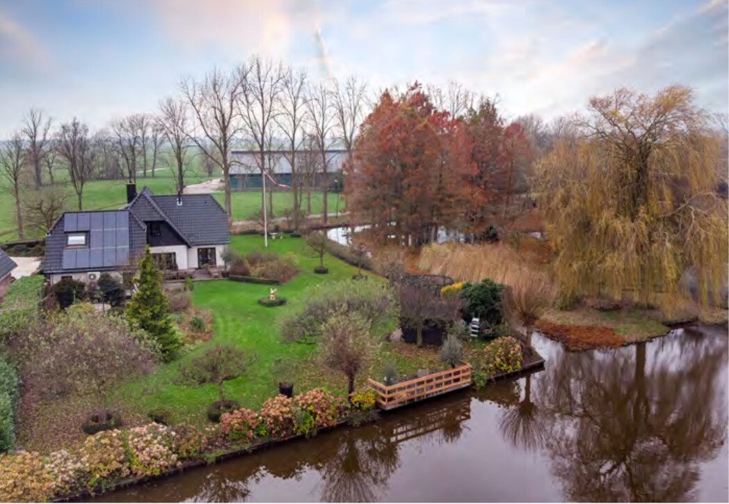 An aerial view of the villa showcases a large, landscaped garden with a variety of trees lining the serene riverbank. The water reflects the surrounding greenery and colorful foliage. A wooden dock is visible at the water's edge, enhancing the outdoor space.