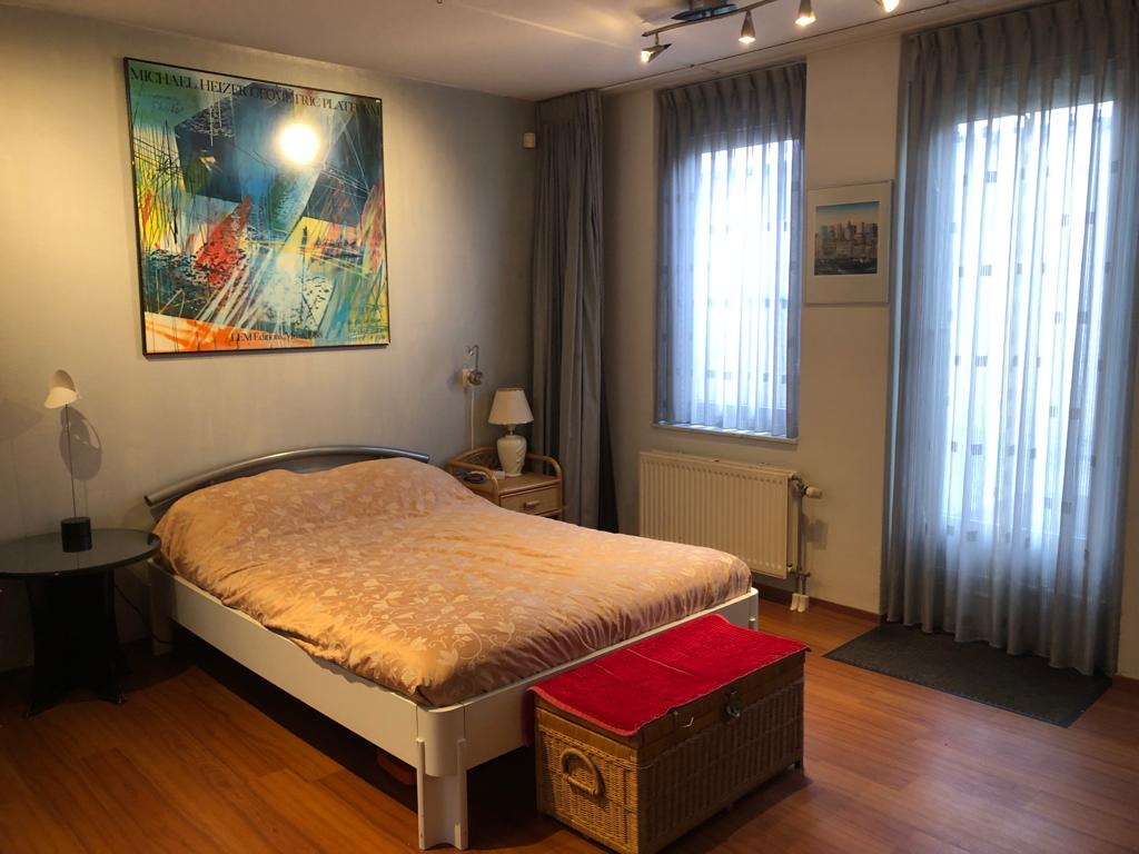 A spacious room is presented, featuring a large bed and warm-toned bedding. Natural light enters through two tall, softly draped windows. The walls are adorned with artwork, and a bedside lamp is positioned on a small table. A wicker storage chest sits at the foot of the bed.