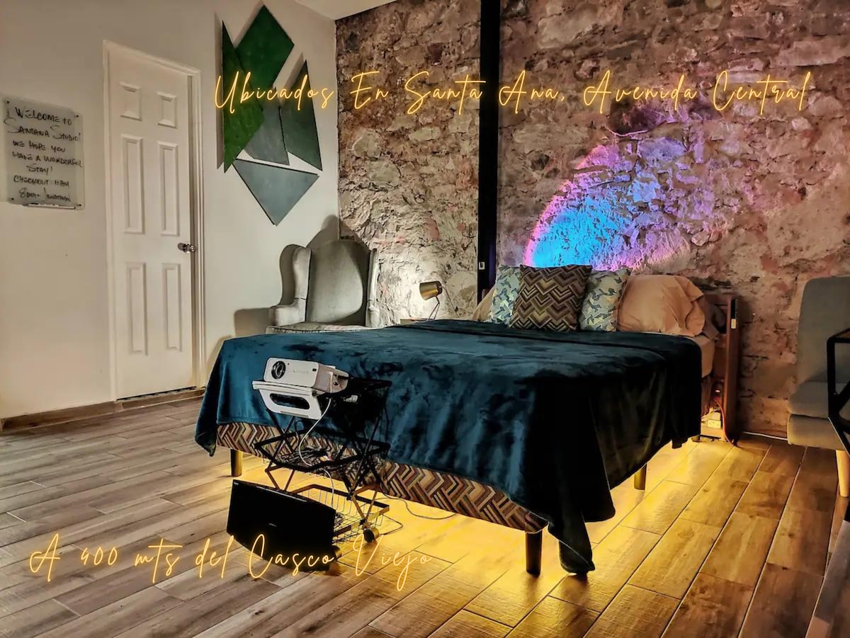 A spacious sleeping area features a queen-size bed with a dark blue coverlet, illuminated subtly by warm lights beneath. A historic stone wall adds texture. A projector and a working desk are positioned nearby, creating a multifunctional space for relaxation and productivity.