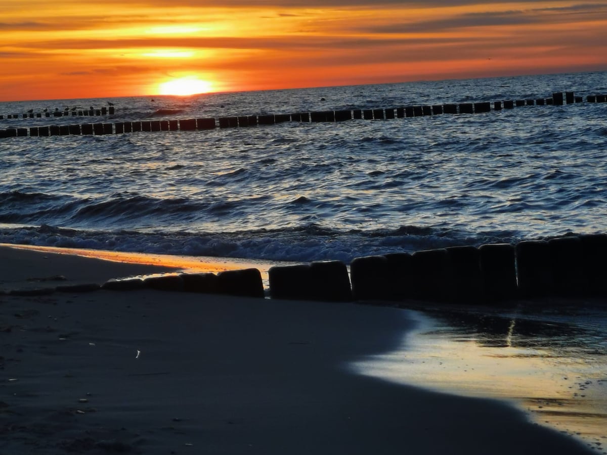 A serene beach scene is captured at sunset, showcasing waves gently lapping at the shore. The horizon displays vibrant colors as the sun sets over the water. Wooden poles extend into the ocean, marking the boundary of the beach.