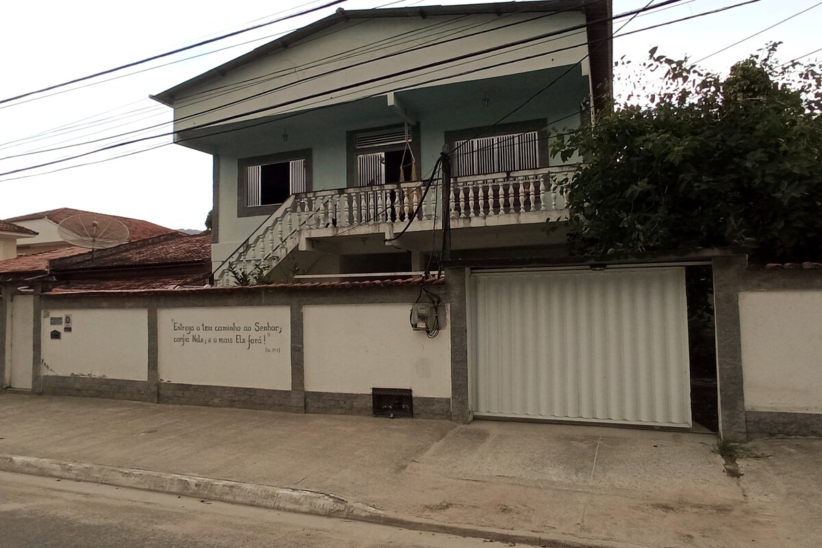 A two-story house presents a simple exterior with a light blue upper level and white railing on the balcony. A white gate is visible in front, leading to a paved area. Nearby, utility lines are seen above the structure.