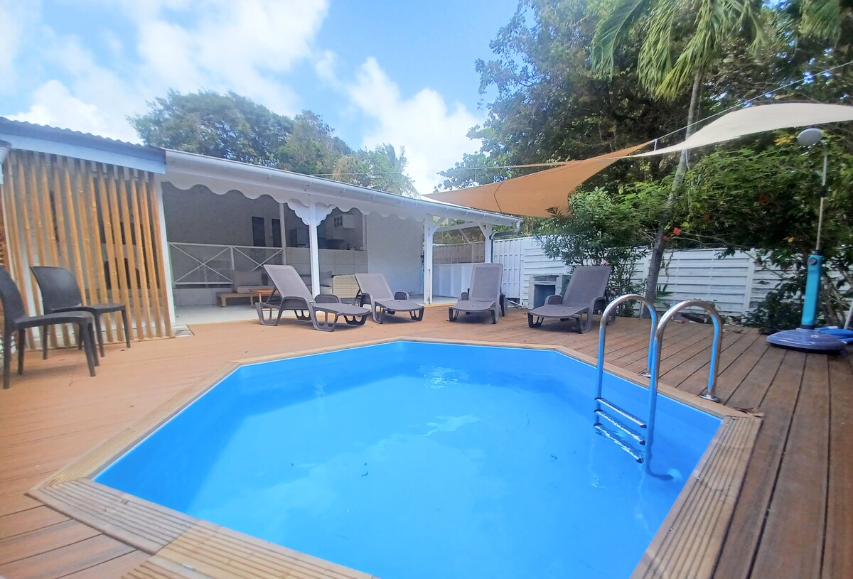 A private, heated pool is surrounded by a spacious wooden deck, featuring several lounge chairs for relaxation. A shaded area is present, providing comfort from the sun. The backdrop includes a light-colored building and greenery, contributing to a serene outdoor space.