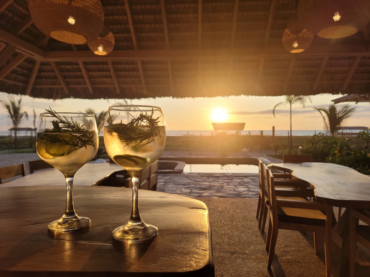 Two elegant cocktails are placed on a wooden table under a thatched roof. The setting sun casts a golden glow over the ocean and pool area in the background, showcasing a serene ambiance complemented by palm trees.