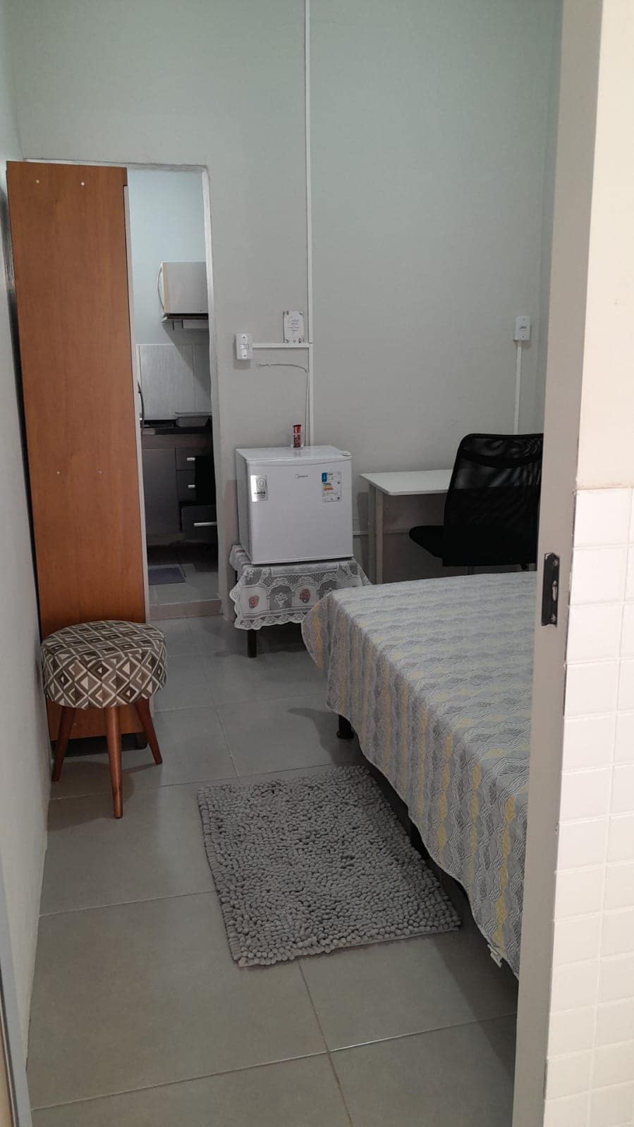 A compact room is displayed, featuring a bed covered with a light-colored patterned blanket. A small table with a chair is positioned nearby, and a mini refrigerator is visible. A rug lies on the tiled floor, contributing to the room's functionality.