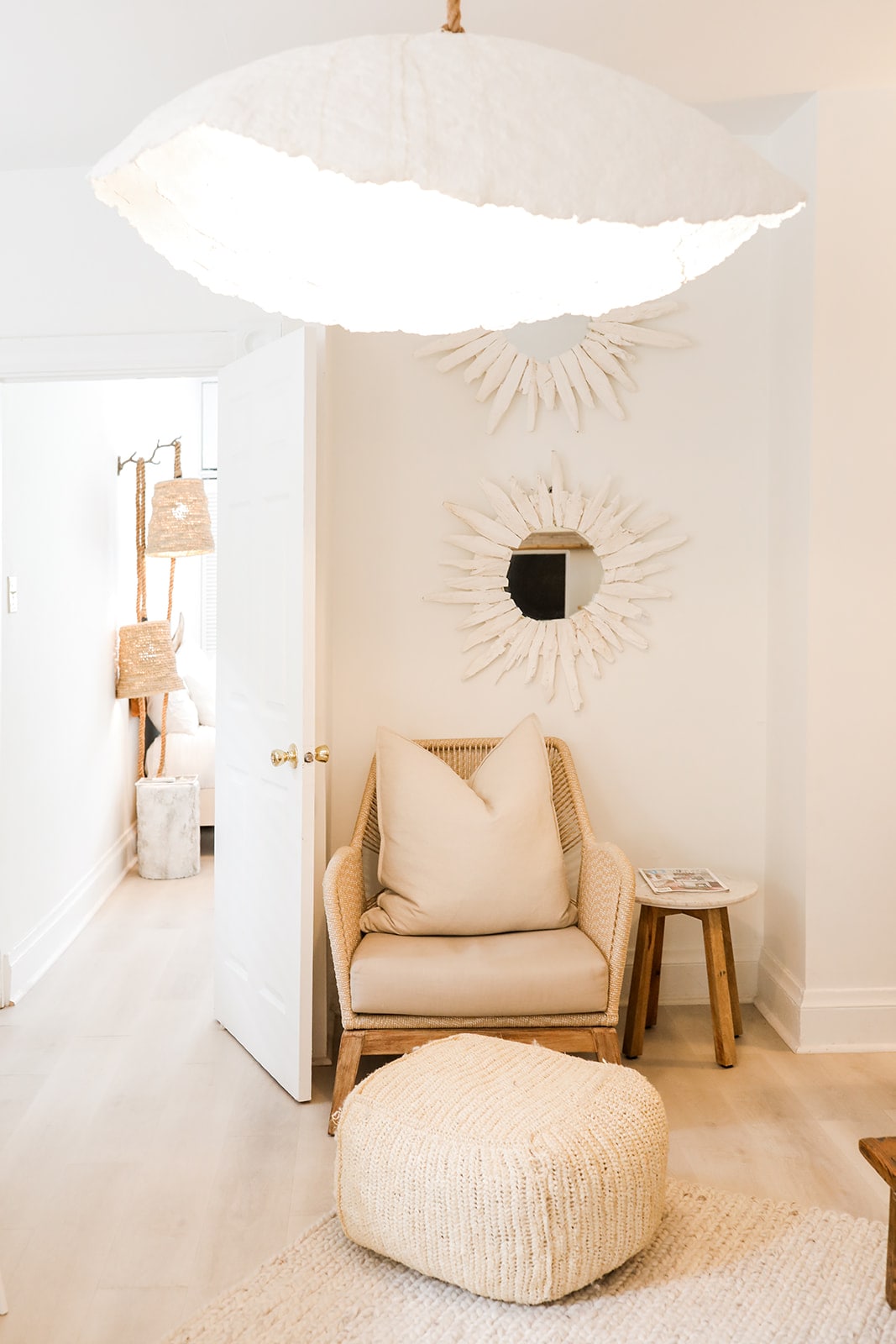 A cozy corner features a rattan chair with a soft cushion, accompanied by a round pouf. A circular mirror hangs on the wall, surrounded by decorative sunburst elements. Soft lighting is provided by a large, textured pendant lamp above.