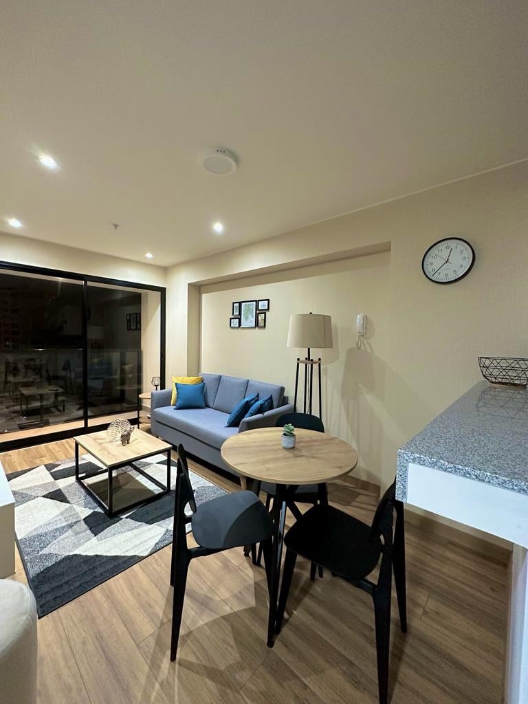 A cozy living area features a comfortable sofa with colorful cushions, accompanied by a small round dining table and four chairs. A modern clock is displayed on the wall, and a large window allows natural light to fill the space, revealing a glimpse of the city.