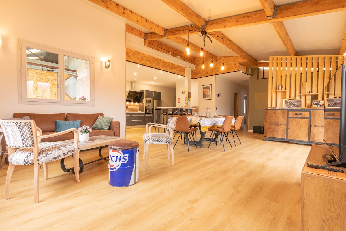 An open living area features wooden beams on the ceiling, a comfortable brown couch, and a round dining table surrounded by chairs. A well-equipped kitchen can be seen in the background, with wooden accents enhancing the space. Natural light filters through the large windows.