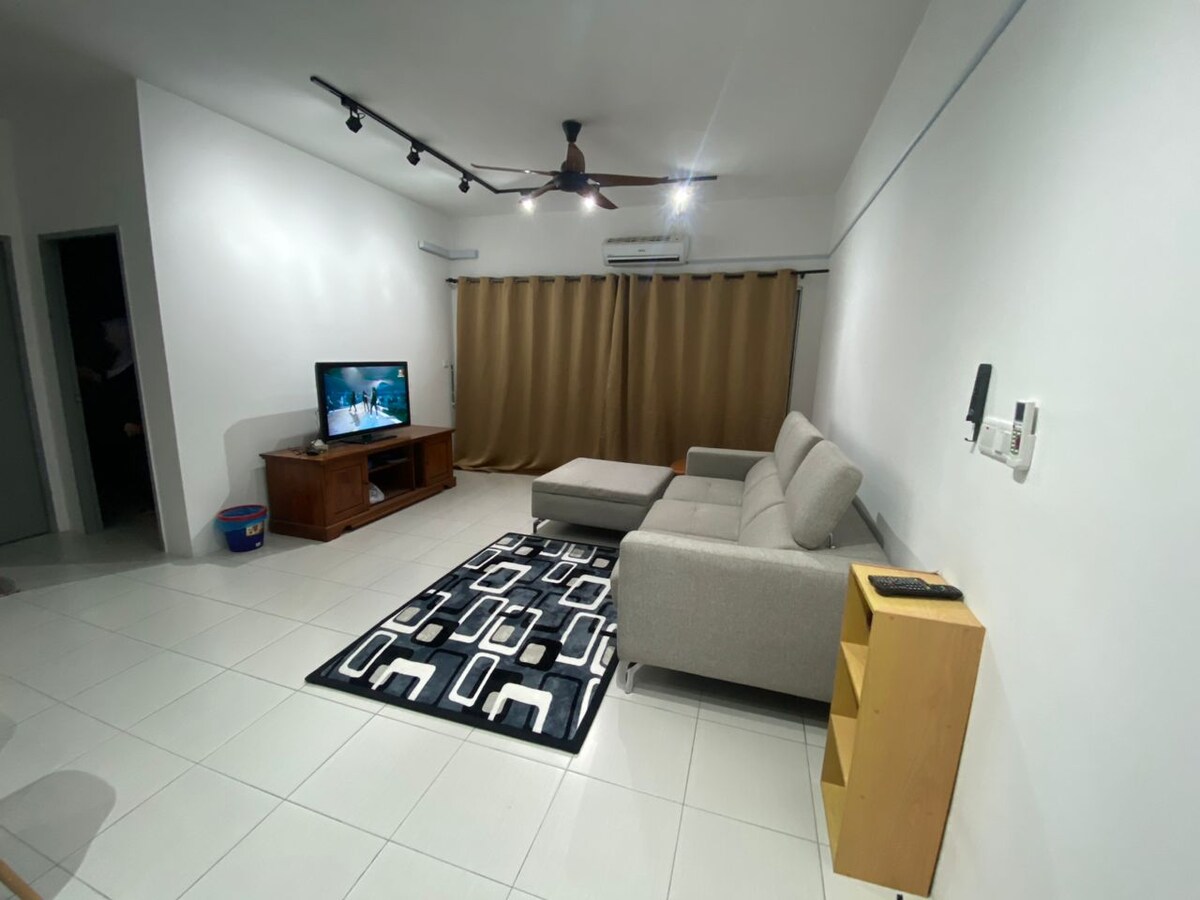 A spacious living area is shown, featuring a comfortable gray sectional sofa and a wooden coffee table. A flat-screen TV is mounted on a wooden media console. Neutral curtains partially cover the windows, allowing soft light to fill the room. A patterned rug adds visual interest.