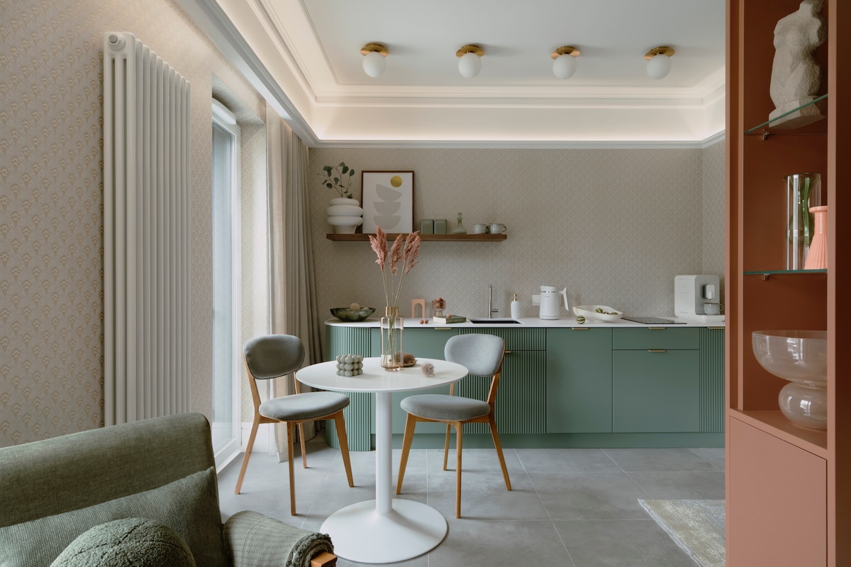 A modern kitchen area features a round white dining table surrounded by three wooden chairs. Green cabinetry lines the wall, complemented by open shelving displaying elegant dishware. Soft lighting enhances the ambiance, while large windows offer natural light, creating a welcoming space.