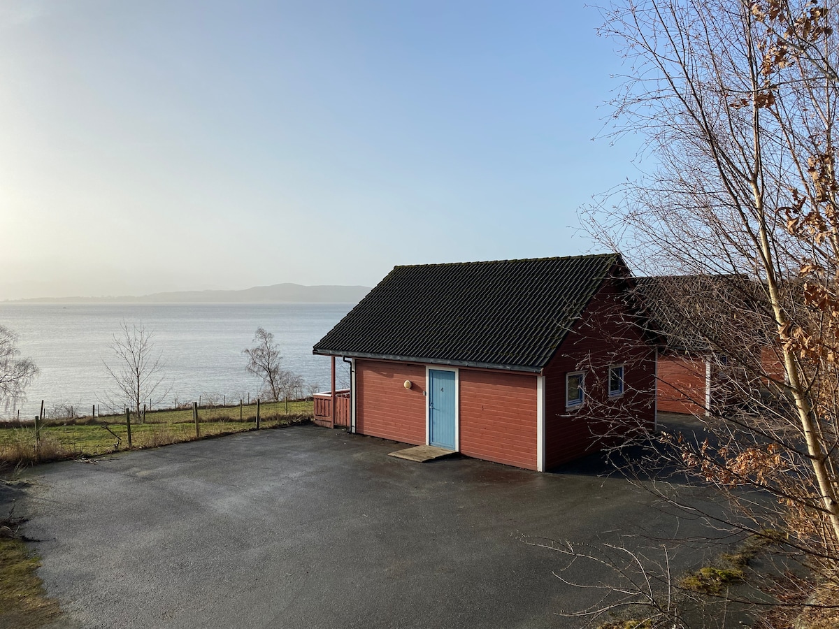 A red cabin with a light blue door is positioned near the water's edge, surrounded by grassy areas and trees. A clear view of the lake and distant hills is visible, while the cabin's entrance is accessed via a paved path.