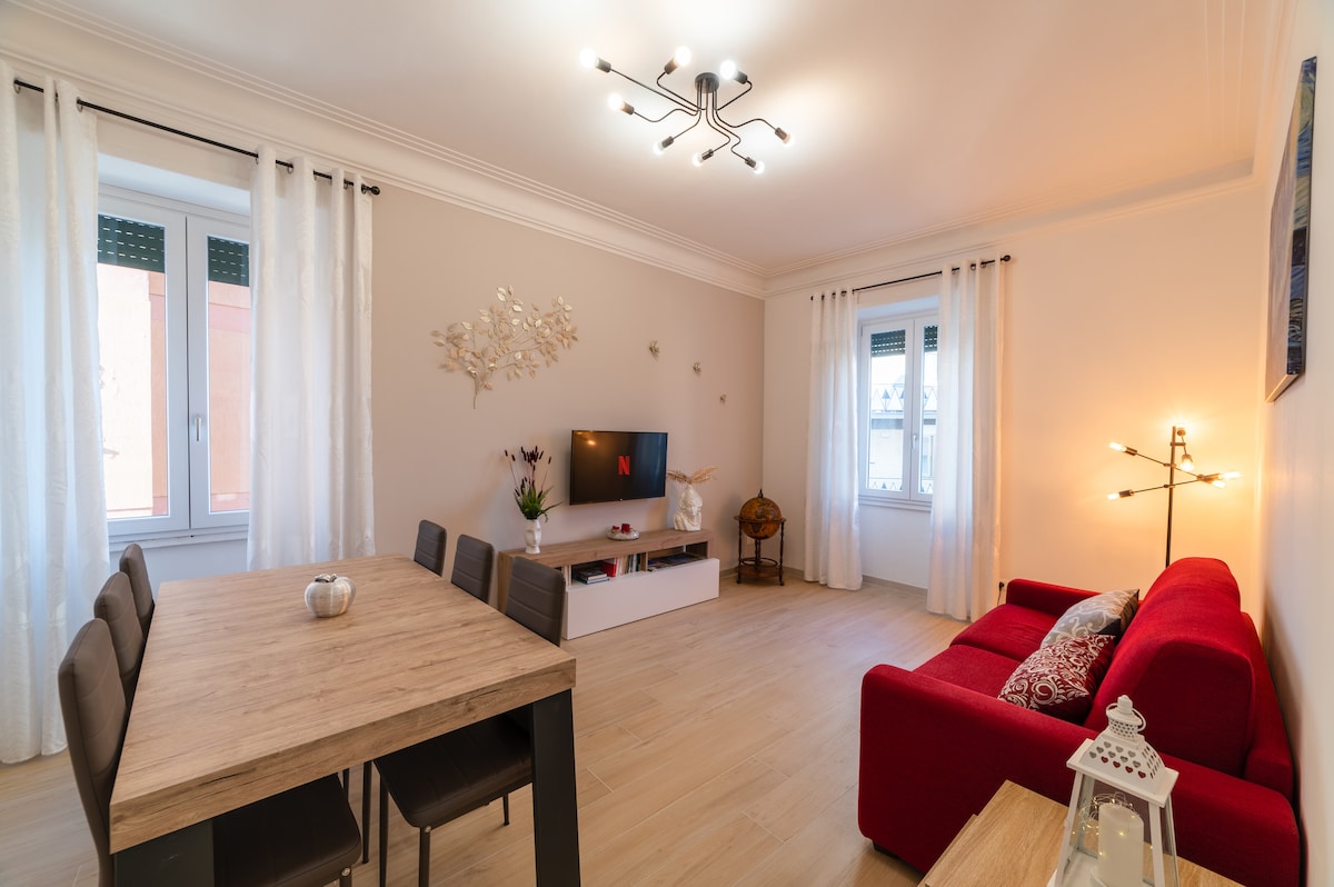 A functional living space features a red sofa paired with a wooden dining table surrounded by black chairs. Natural light enters through two windows dressed with white curtains. A TV is positioned on a low cabinet, with decorative elements enhancing the room's simplicity.