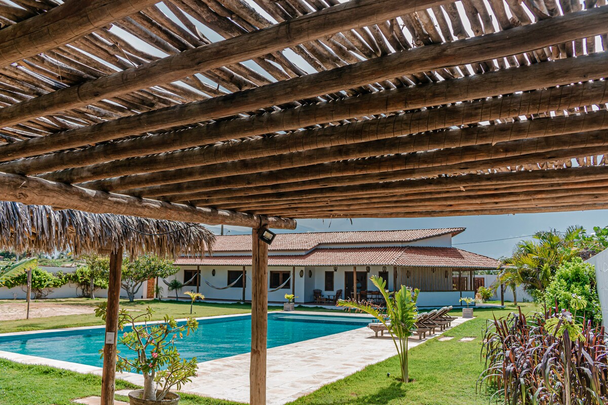 A spacious outdoor area reveals a large swimming pool surrounded by verdant landscaping. The structure features a wooden pergola overhead, providing shade. Loungers are positioned near the pool, while the white building in the background offers a relaxed, inviting space for gatherings.
