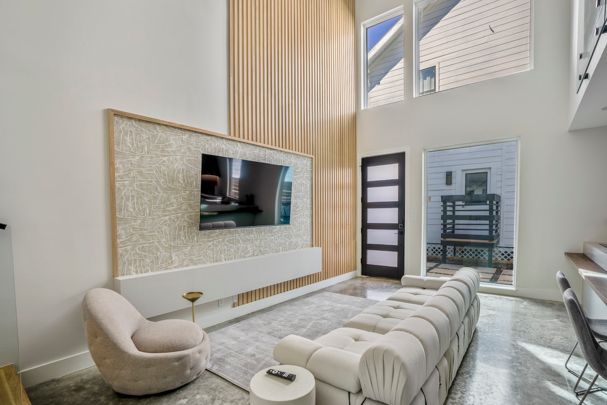 A bright living area is characterized by tall ceilings and large windows, allowing natural light to fill the space. A plush sectional sofa faces a wall-mounted television, while a sleek side table stands nearby. Light neutral tones emphasize a modern design.