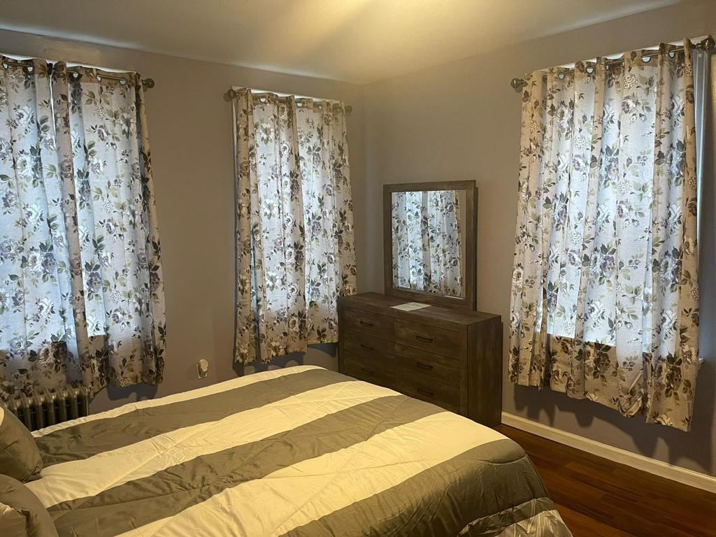 A cozy bedroom features a queen bed with a striped comforter. Natural light enters through two floral-patterned curtains adorning the windows. A wooden dresser with a matching mirror complements the soft, neutral wall color and warm hardwood flooring.
