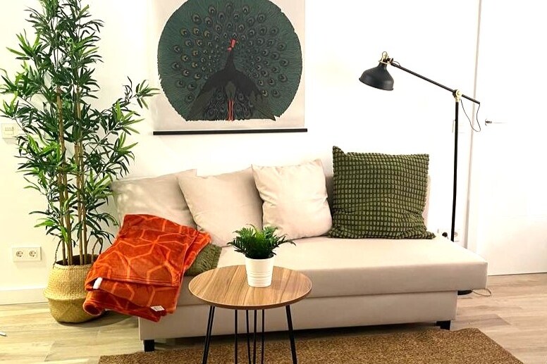 A cozy seating area is presented with a light-colored sofa, adorned with green and orange cushions. A small round table sits in front, accompanied by a decorative plant. A vibrant peacock artwork adds color to the wall, while a floor lamp provides additional lighting.