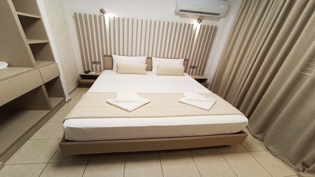 A well-lit sleeping area features a spacious bed with neutral-toned bedding, centered against a modern panelled headboard. Soft lighting is provided by wall-mounted fixtures. The room includes built-in shelving and light-colored curtains that frame a window.