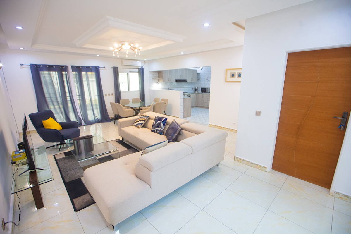 A spacious living area is filled with natural light, featuring a neutral-colored sectional sofa and a dark coffee table. Stylish chairs surround the dining area, and a modern kitchen is visible in the background. Large windows are dressed in dark curtains, providing a serene atmosphere.