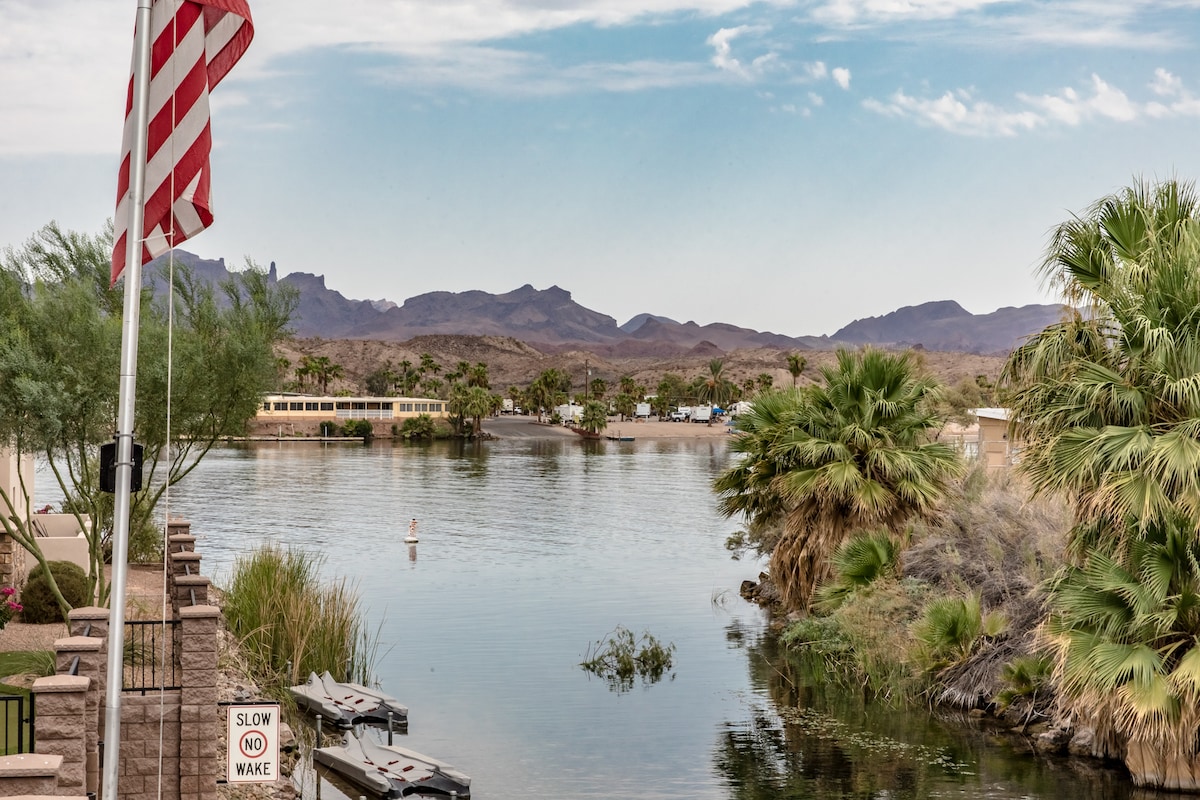 A serene view of a calm canal is framed by tall palm trees and lush greenery. In the distance, gentle hills provide a backdrop under a clear sky. An American flag stands along the water's edge, signaling the peaceful atmosphere of the surroundings.