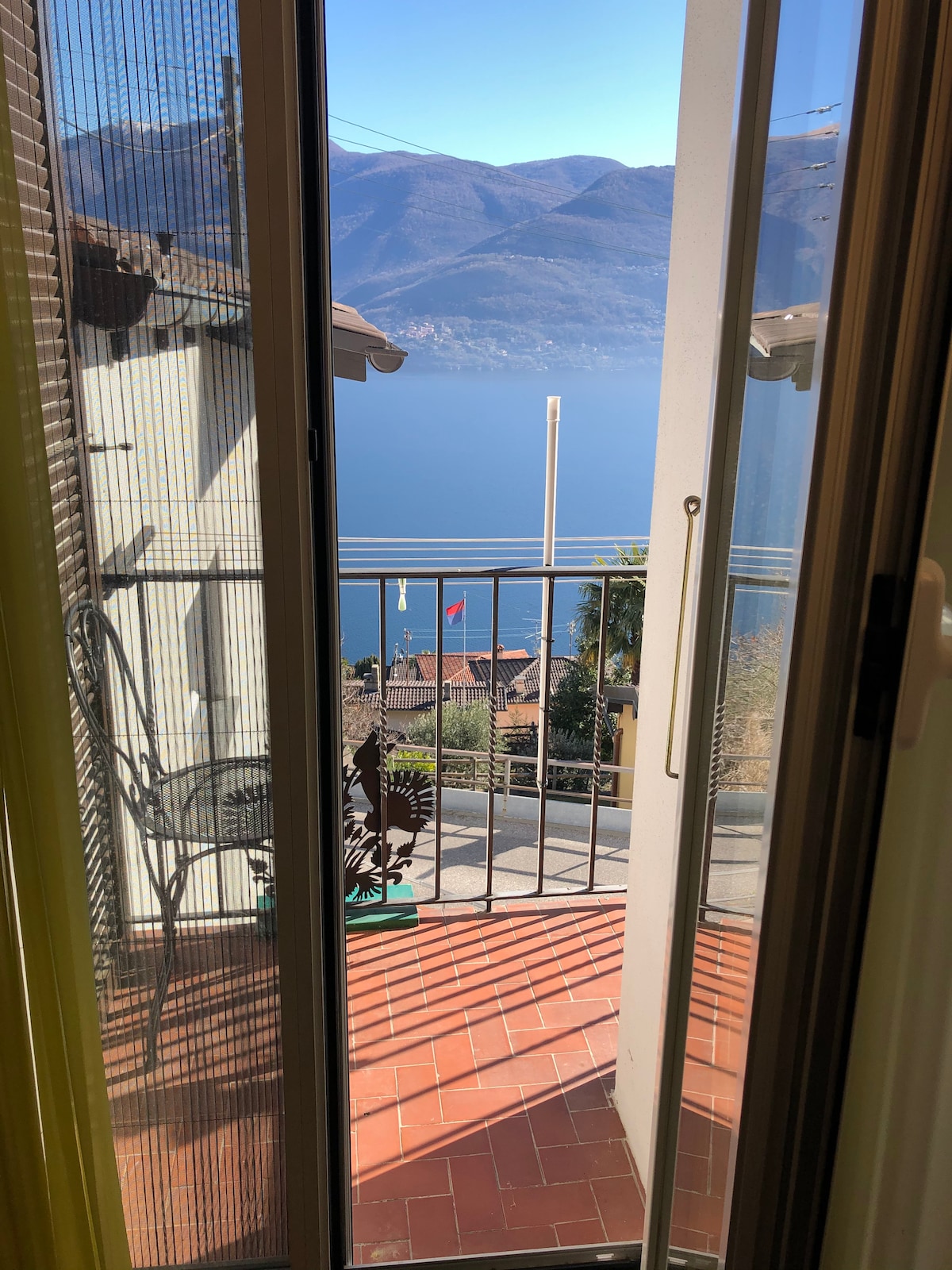 A doorway opens to a balcony featuring a metal railing, providing a view of the serene Lago Maggiore and surrounding mountains. The flooring is tiled, and a small chair is positioned to enjoy the landscape.