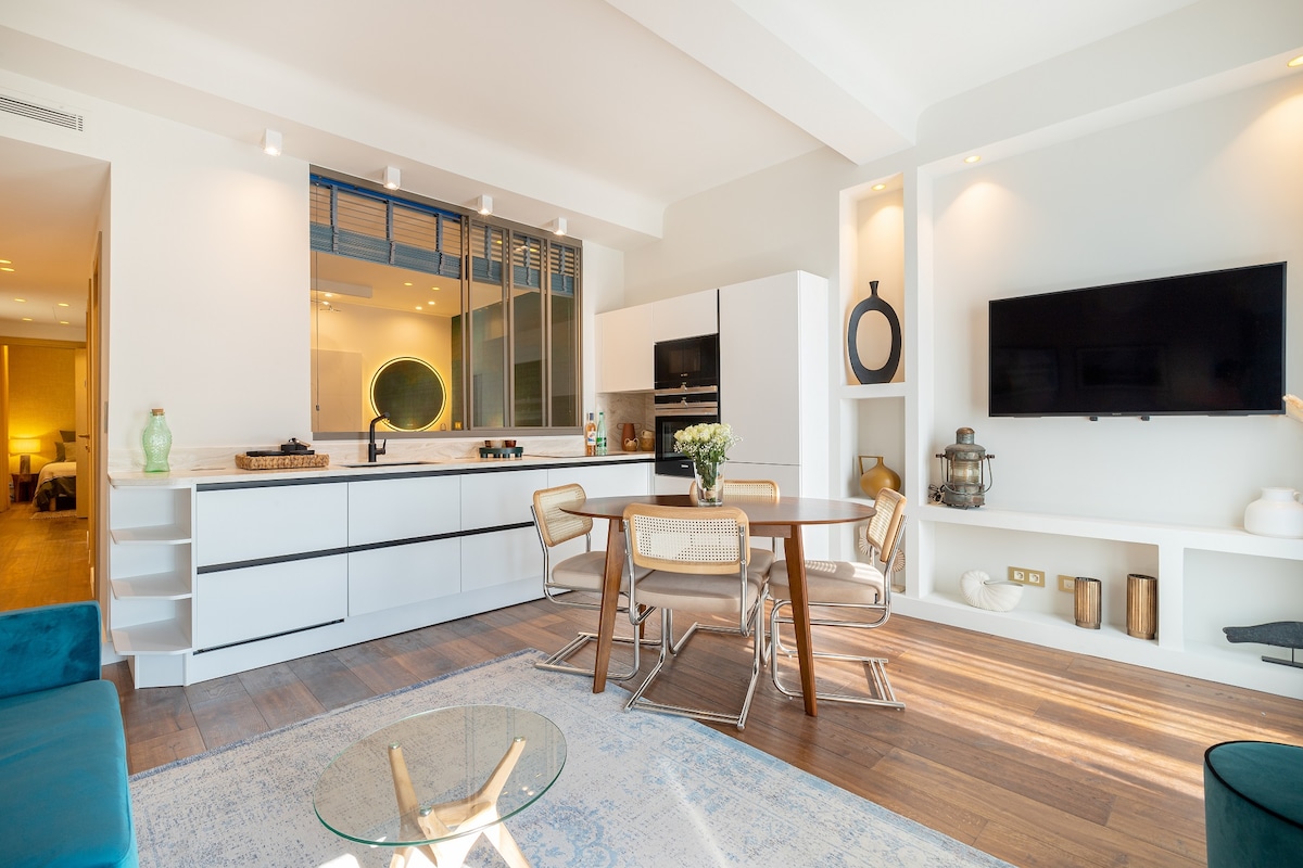 A modern kitchen and living area is showcased with sleek white cabinetry and a polished wooden dining table surrounded by stylish chairs. Natural light floods the space, highlighting the elegant design features, including a wall-mounted smart TV and decorative accents on a console.