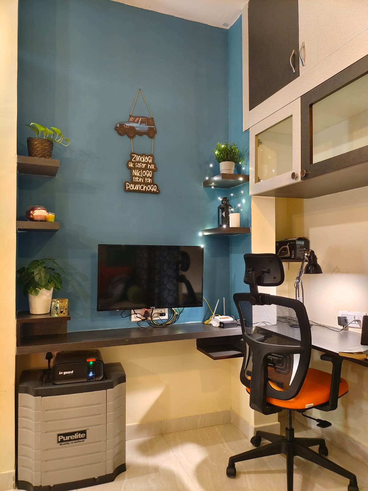 A compact workspace is shown with a wall-mounted desk and ergonomic chair. A television is positioned against the wall. Stylish shelves display decorative plants and ambient lighting. The space features a mix of blue and neutral tones, creating a calming and functional environment.