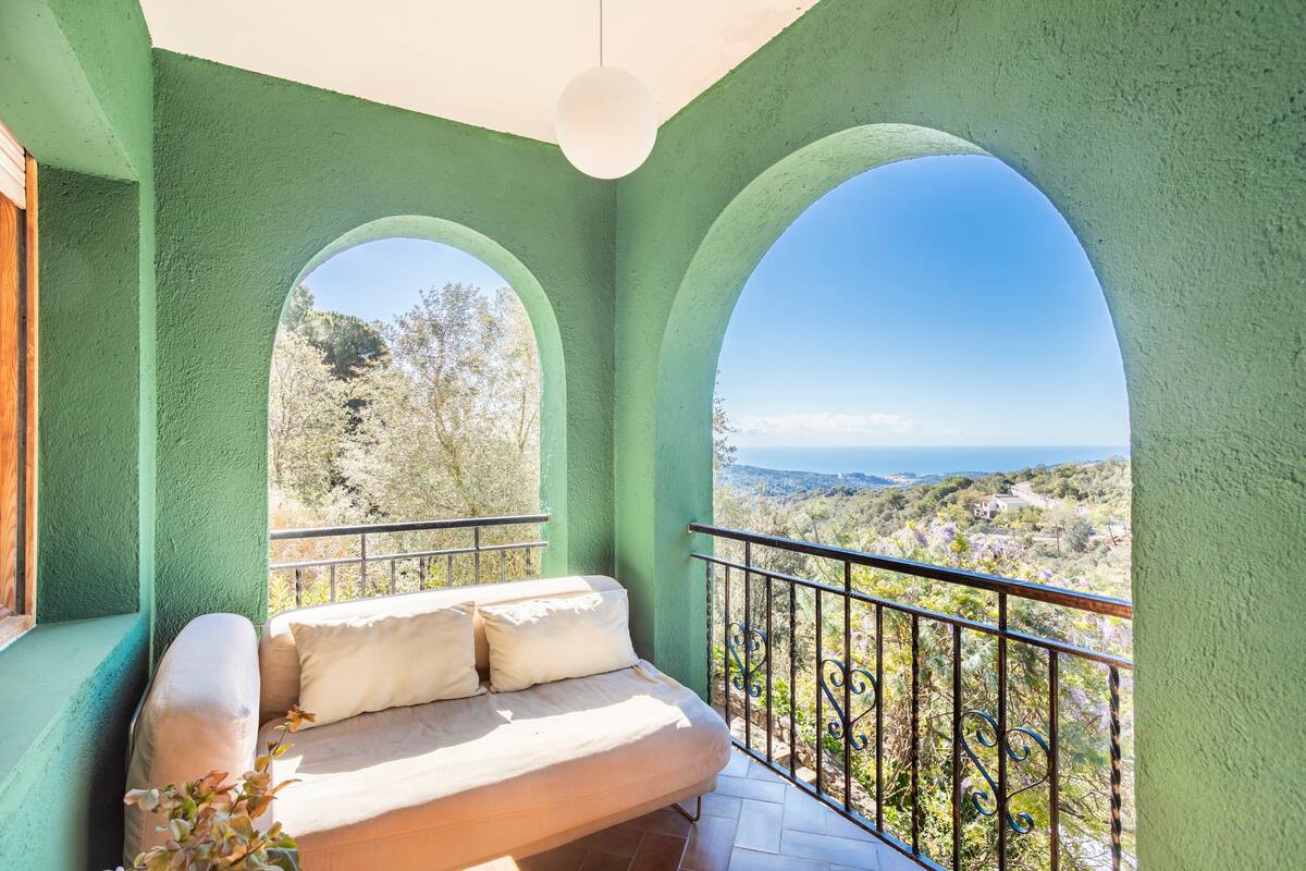 An outdoor seating area is framed by two archways, offering views of the expansive landscape and the sea. A light-colored sofa rests against the wall, complemented by a pendant light overhead. The vibrant green walls enhance the connection with nature.