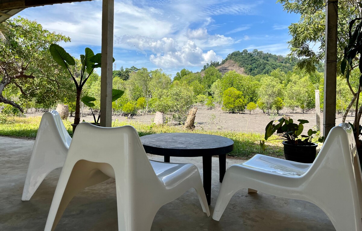 An outdoor seating area is visible, featuring two white chairs and a round black table. Lush greenery and mangrove trees are seen in the background, with a clear blue sky overhead. The tranquil surroundings promote a peaceful atmosphere.