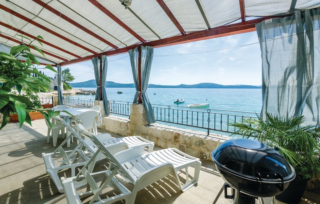A spacious terrace is shown with a partial view of the sea, featuring lounge chairs and a grill. Sunlight filters through the draped canopies, while lush green plants add a touch of nature to the outdoor space. Calm waters and distant hills create a serene backdrop.